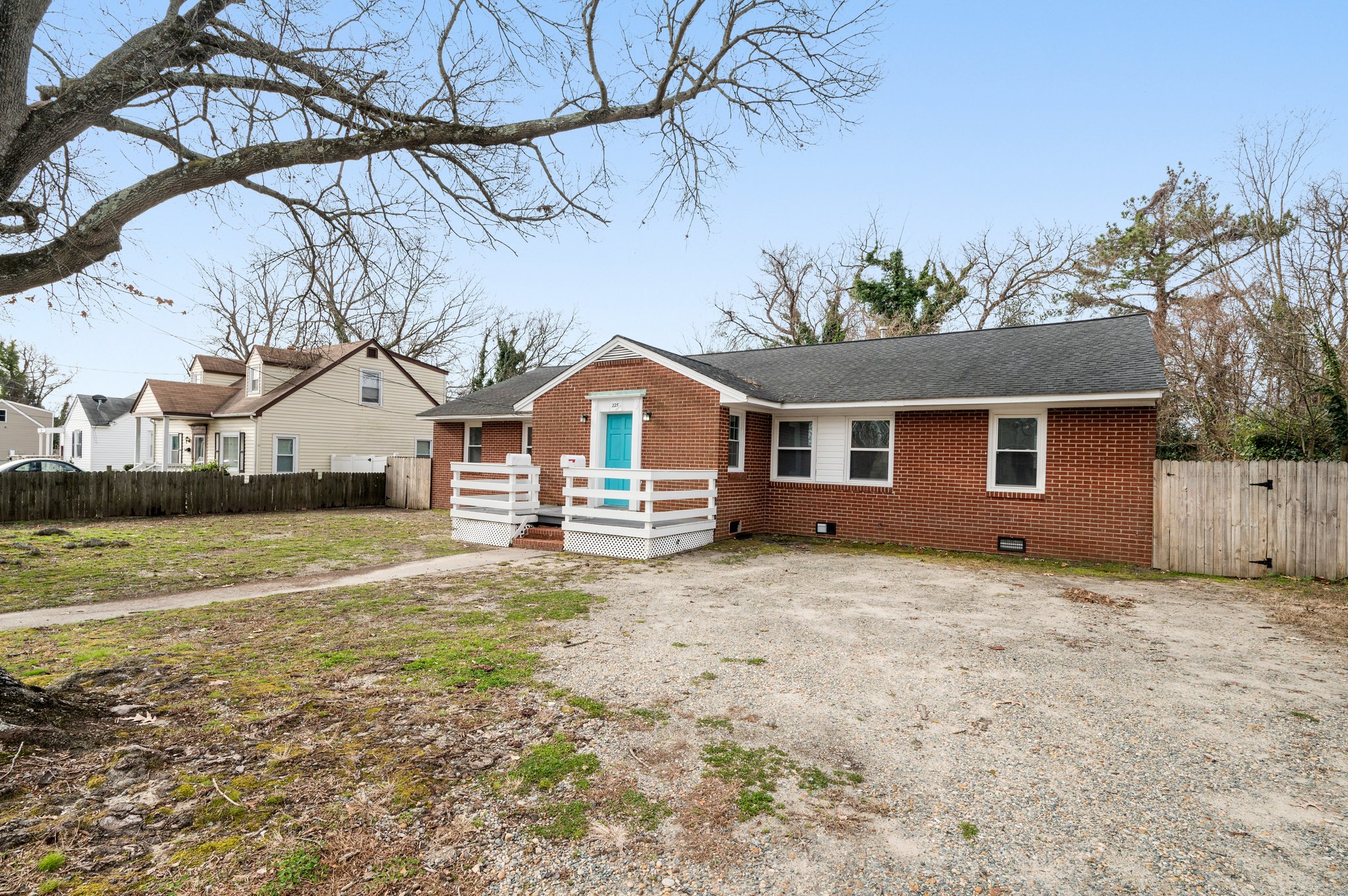 Angled front view of a red brick duplex with a gravel driveway and fenced backyard.