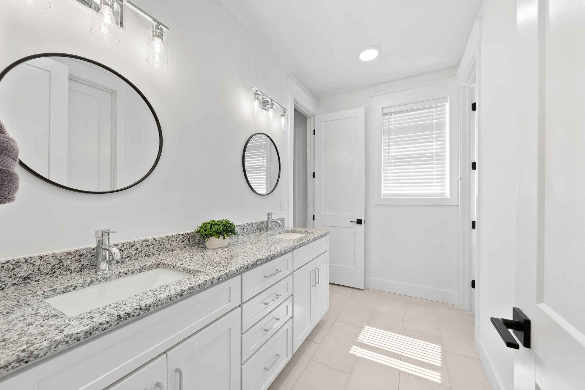 A bright white bathroom with a granite double vanity, black-framed round mirrors, and modern light fixtures.