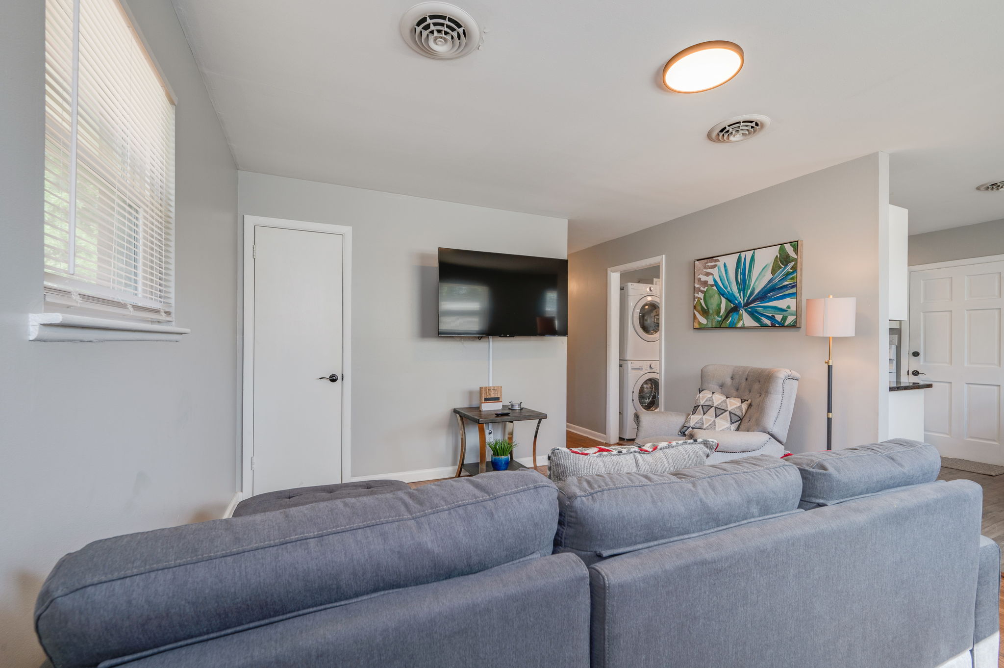 Living room with a gray sectional sofa, mounted flat-screen TV, and a washer-dryer in the background.