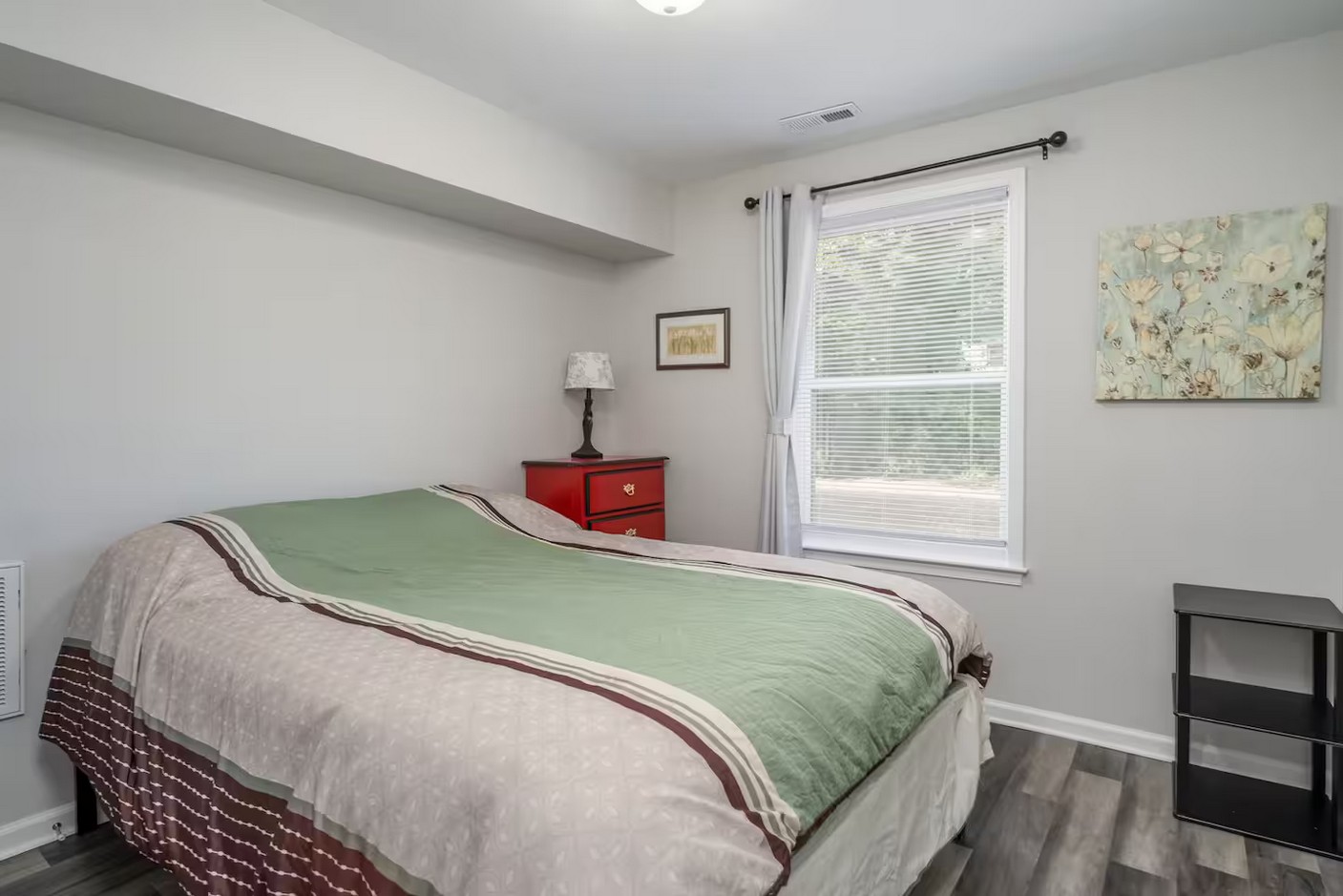 Cozy bedroom with green bedspread, red nightstand, and large window with white curtains.