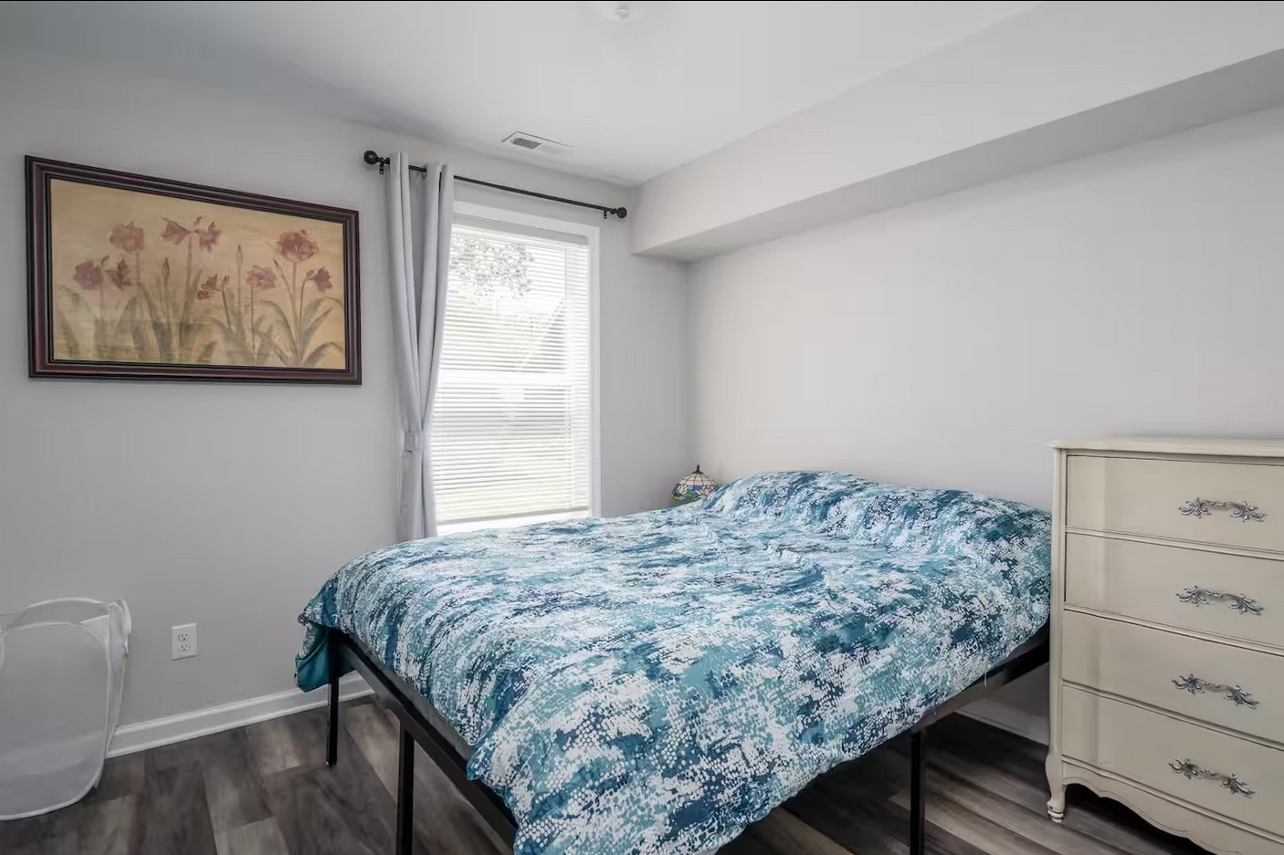 Bright bedroom featuring a blue bedspread, white dresser, and a large window with blinds.