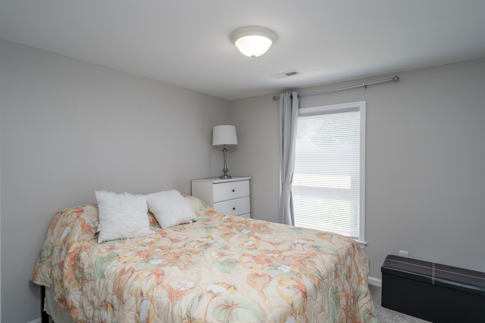 Bright bedroom with a floral bedspread, white dresser, and window with curtains.