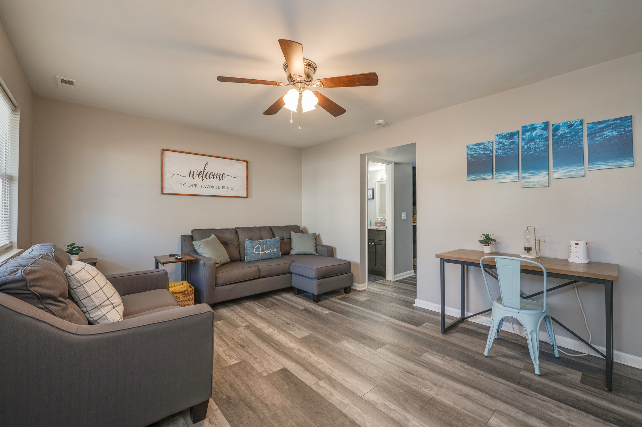 Living room with gray sofas, a wooden desk with a light blue chair, wall art, and a ceiling fan with lights.