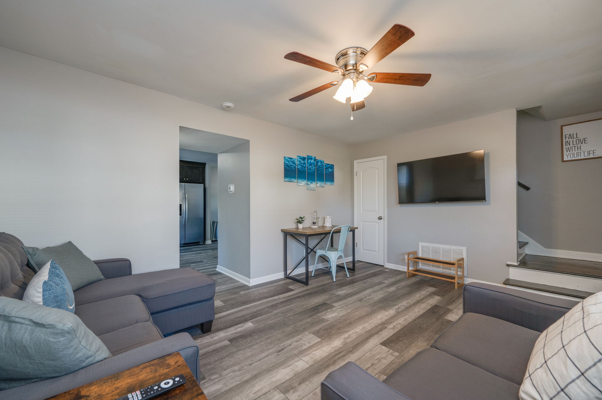 Living room with gray sectional sofa, wall-mounted TV, desk with chair, and view into the kitchen.