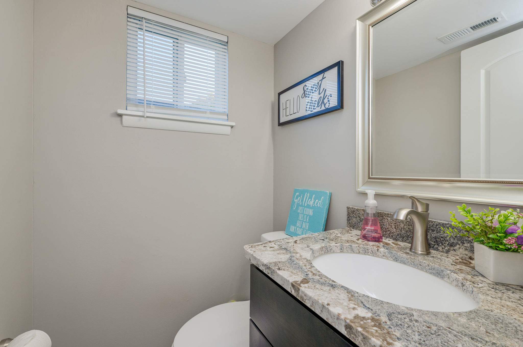 Bathroom with granite countertop, white sink, mirror, small window, and decorative wall signs.