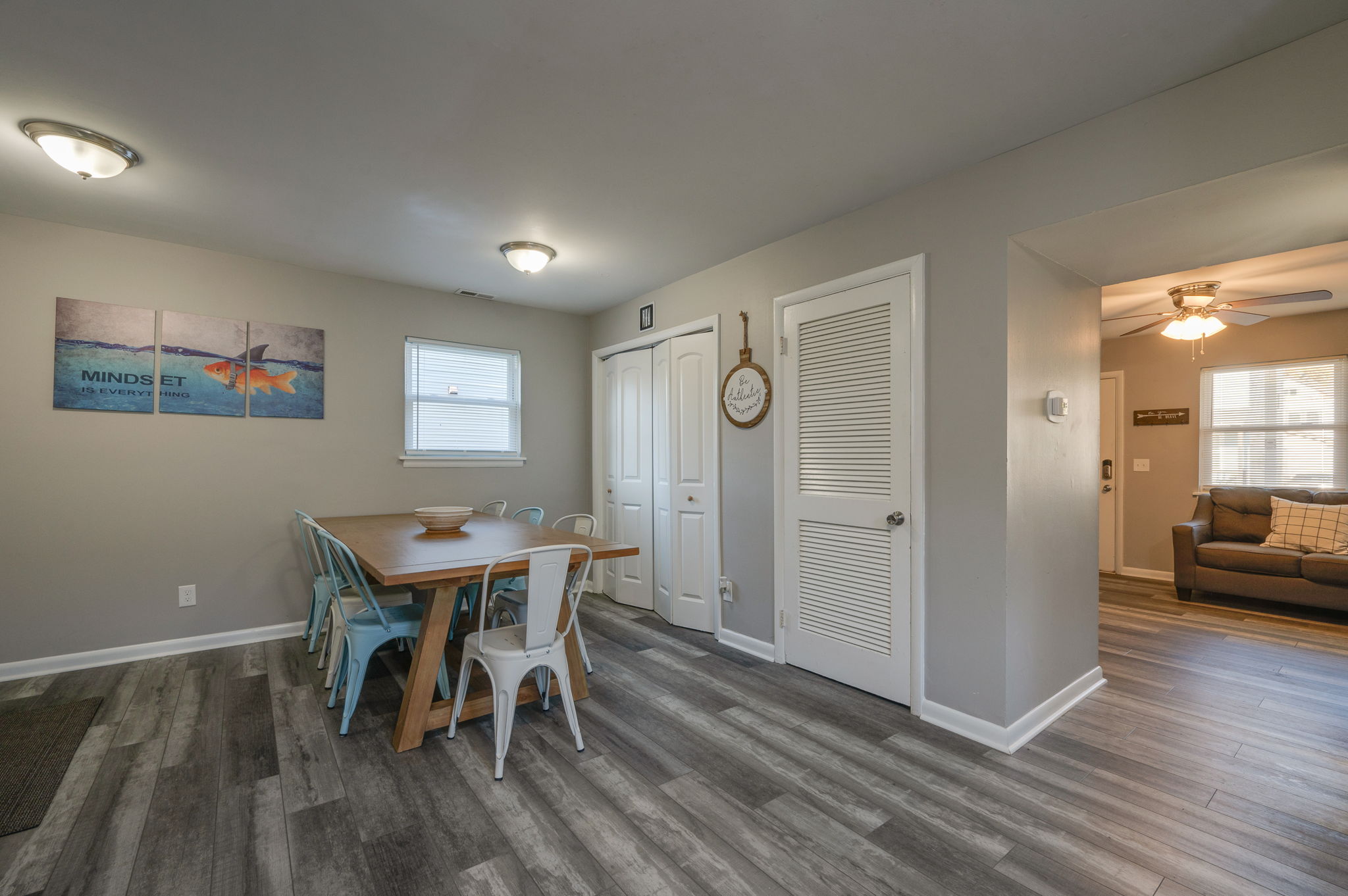 Dining area with wooden table, light blue metal chairs, and wall art of a fish with 'Mindset is Everything' text.