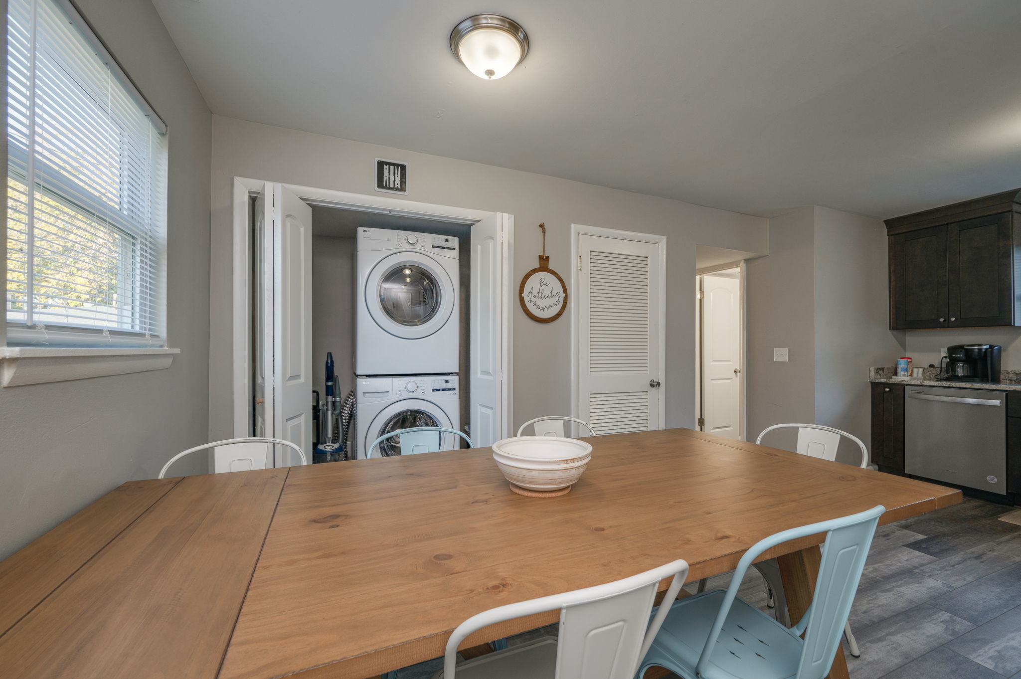 Dining area with wooden table, light blue chairs, and open closet revealing stacked washer and dryer.