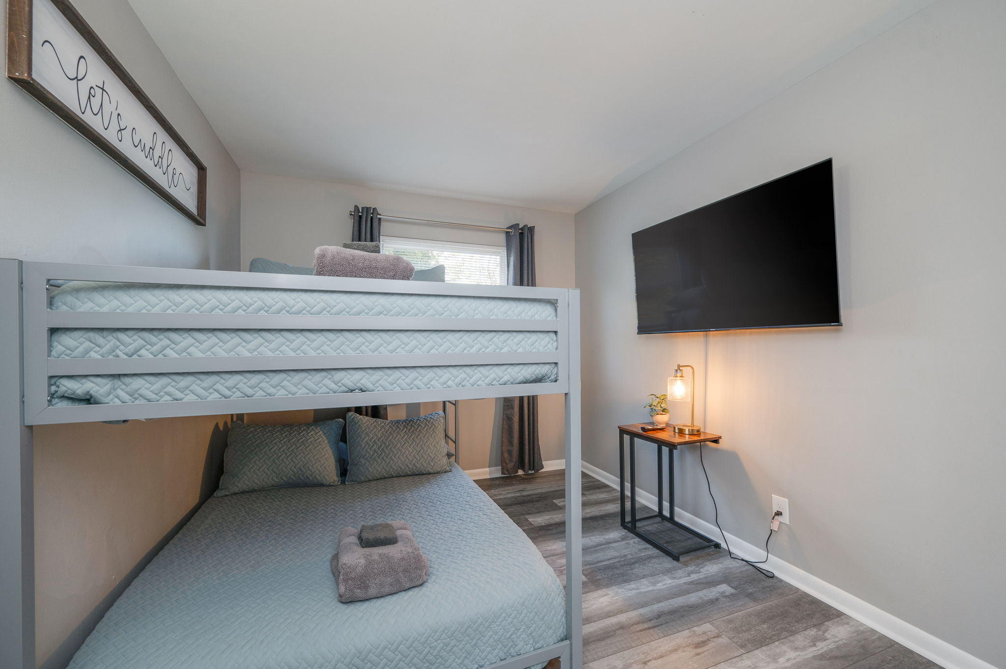 Bedroom with light blue bunk bed, towels on bed, wall-mounted TV, and side table with lamp.