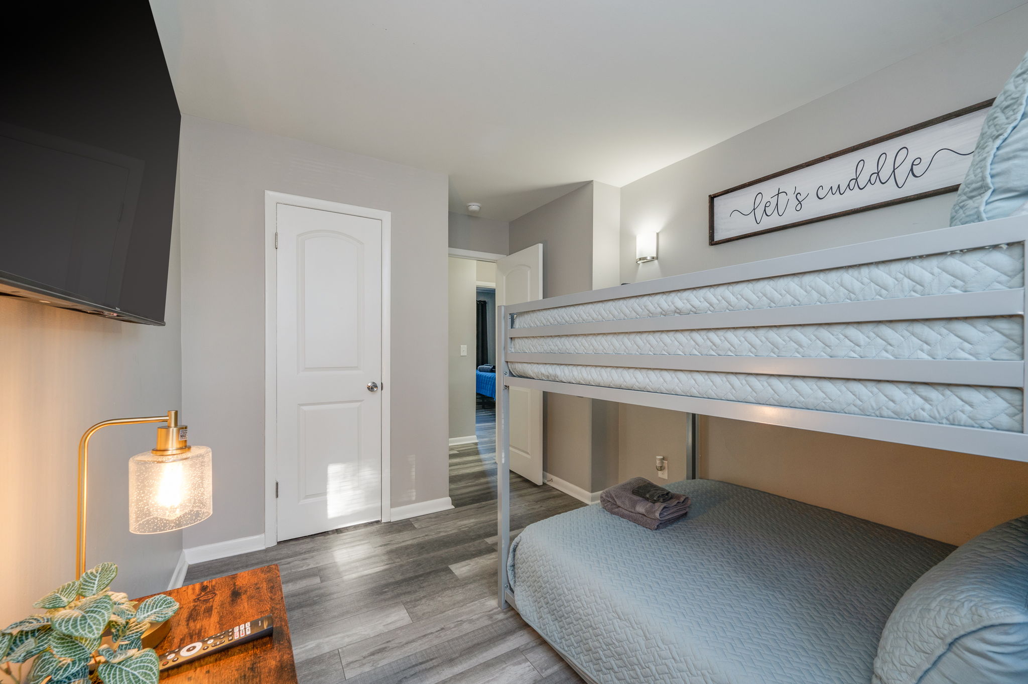 Bedroom view showing light blue bunk bed, side table with lamp, and wall art saying 'Let's Cuddle.'