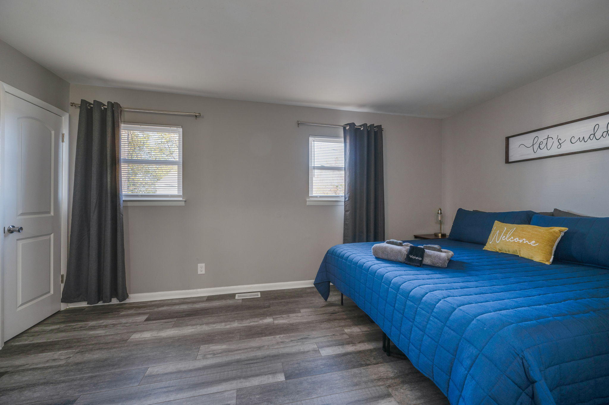Bedroom with large blue bed, yellow 'Welcome' pillow, and two windows with dark curtains.