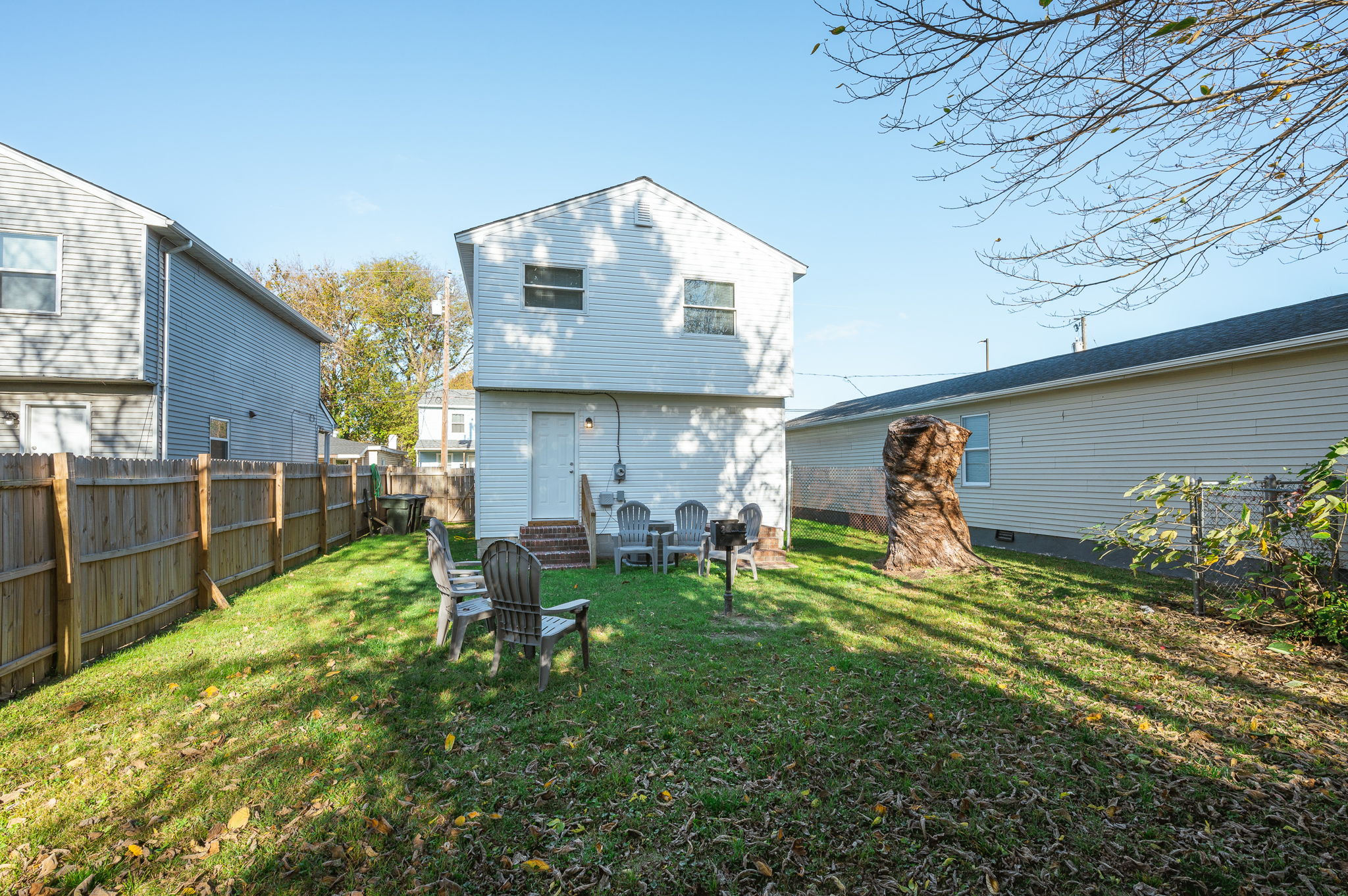Backyard view of white house with fenced yard, outdoor seating, grill, and trimmed tree stump.