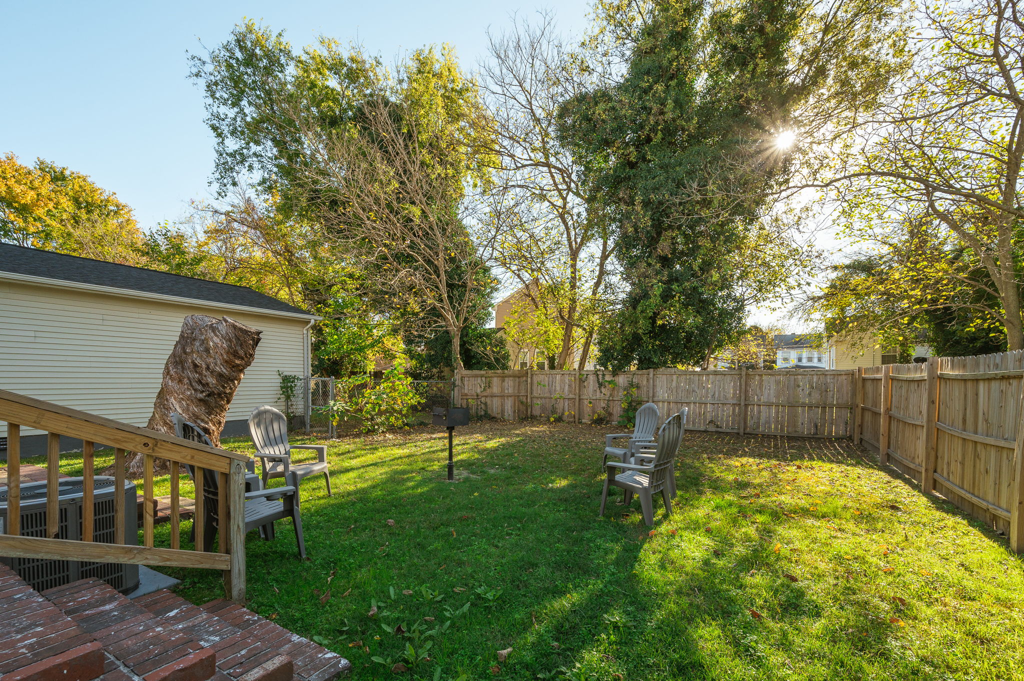 Fenced backyard with green lawn, outdoor seating, and large trees providing shade.